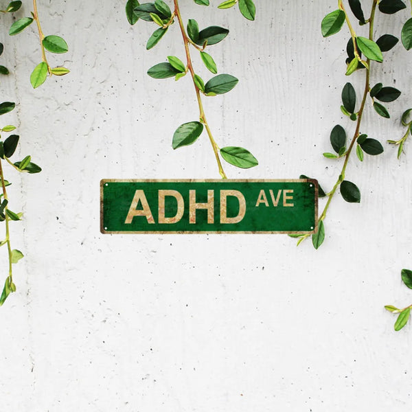 A weathered green metal sign with the text ADHD AVE mounted on a white wall surrounded by green vines.