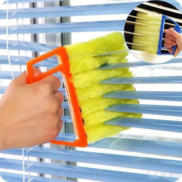 A hand uses an orange multi-functional cleaning brush to clean the slats of blue window blinds.