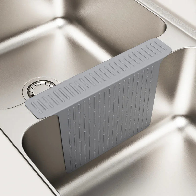 A gray silicone kitchen sink protector mat with drainage features is shown inside a stainless steel sink.