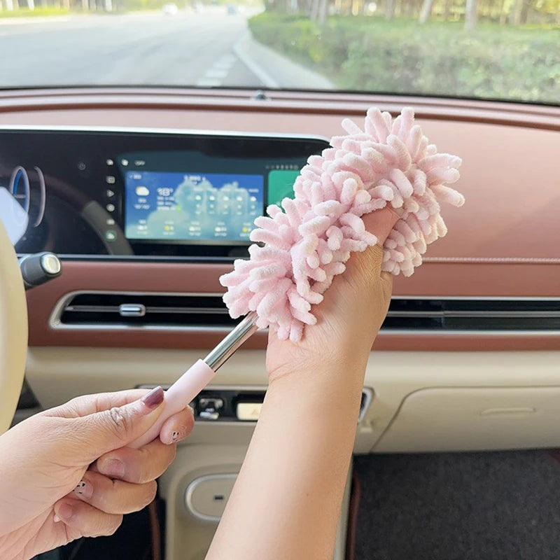 A hand holds a pink extendable microfiber duster brush with fluffy fibers, positioned near a car's interior dashboard.