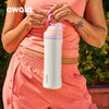A person holds an Owala insulated water bottle in a light pink color against a green background.
