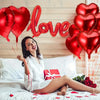 Lächelnde Frau liegt auf dem Bett umgeben von roten Herzballons und Herzkonfetti.