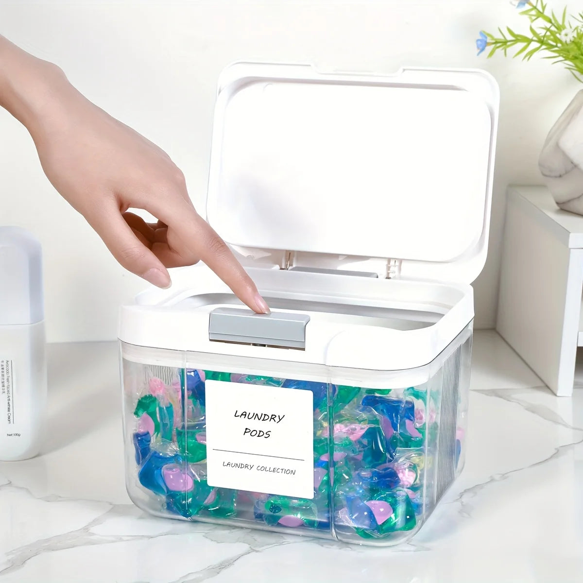 A hand opens a clear plastic laundry pod organizer with a lid, showing colorful laundry pods inside.