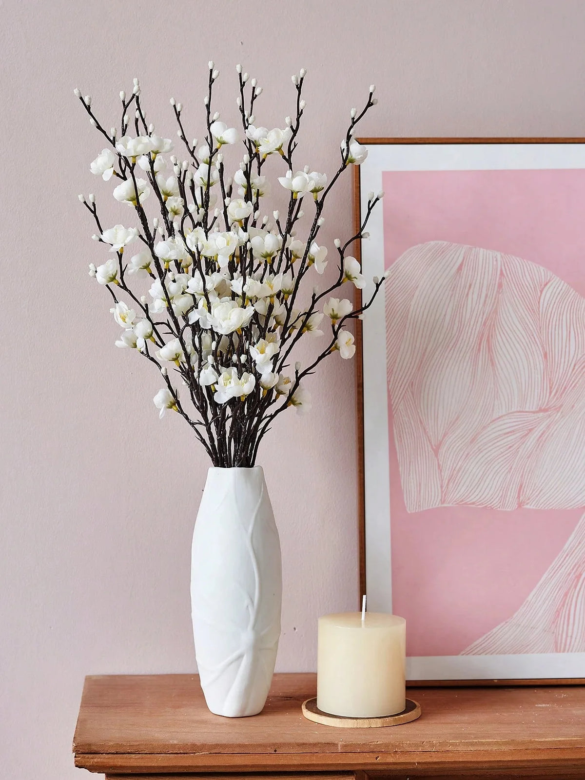 Eine Vase mit künstlichen weißen Pflaumenblütenzweigen auf einer Holzoberfläche neben einem rosa gerahmten Kunstwerk und eine