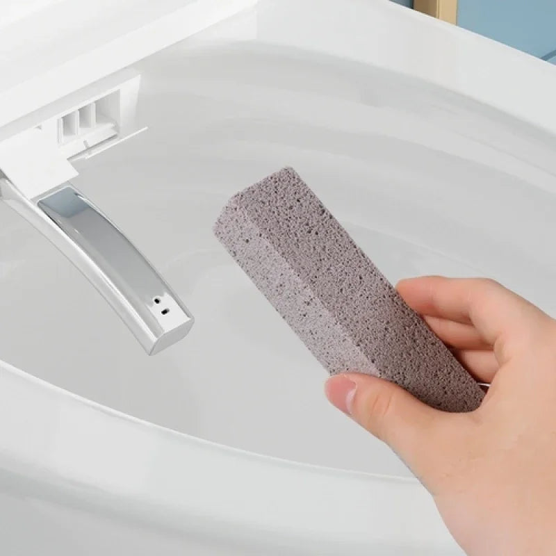 A hand holds a gray pumice stone cleaning stick inside a white toilet bowl for stain removal.