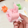 A hand holds a pink anti-splash faucet extender shaped like a cute rabbit against a white background.