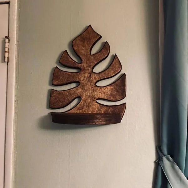 Un cadre d'art mural en bois en forme de feuille de monstera fixé sur un mur vert clair.