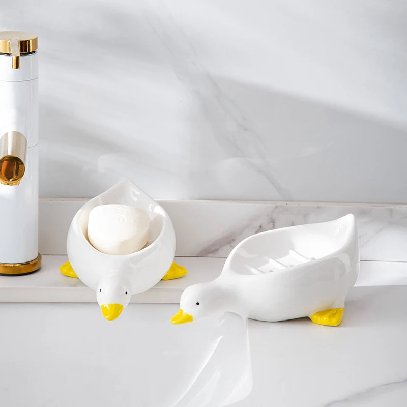 A white duck-shaped soap dish sits on a marble surface next to a gold dispenser.