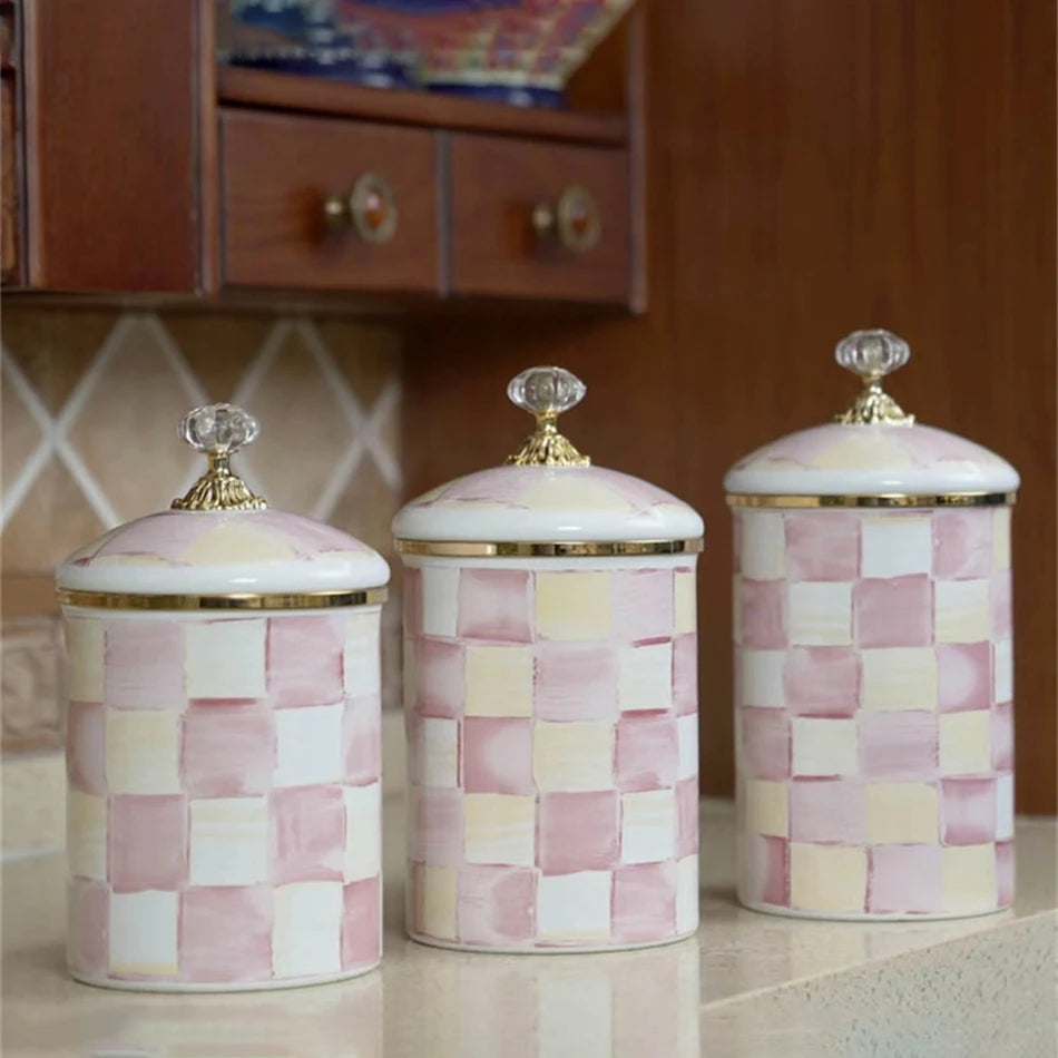 Pink and cream checkered enamel storage jars with gold lids on a light surface.