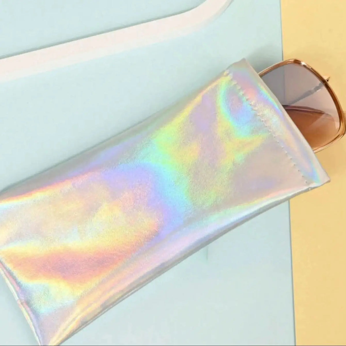 A translucent iridescent PU leather squeeze case for eyeglasses resting on a light blue surface next to sunglasses.