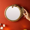 A hand is holding a round, ornate golden mirror frame against an orange wall.
