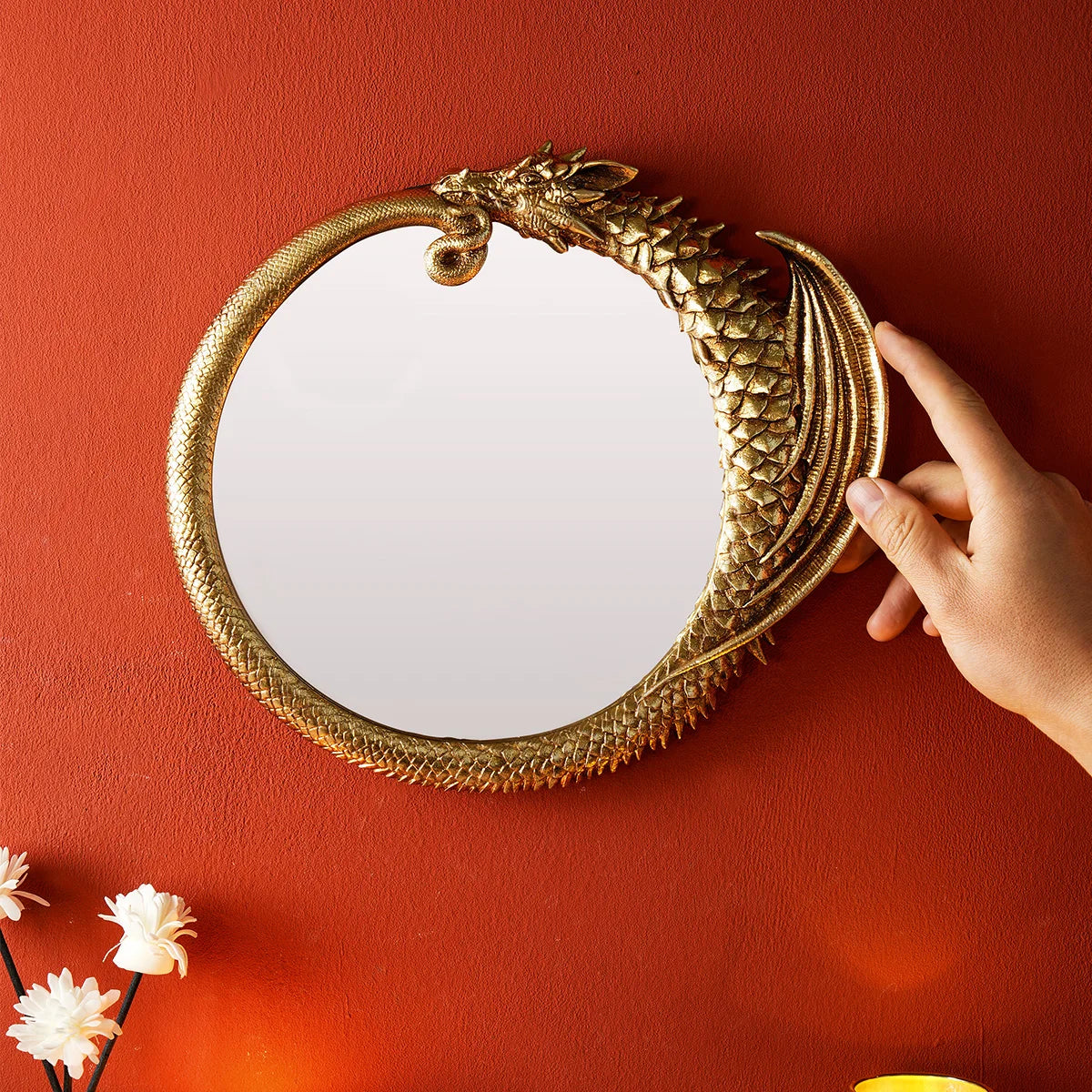 A hand is holding a round, ornate golden mirror frame against an orange wall.