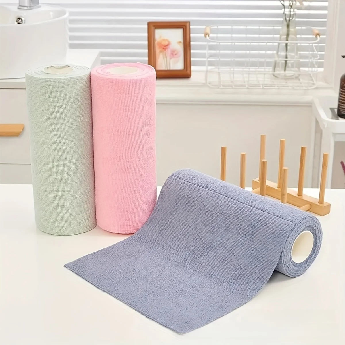 Three rolls of pink and green reusable multi-surface cleaning wipes next to a purple cleaning cloth on a white countertop.