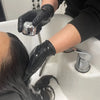 A person wearing black disposable nitrile cleaning gloves is using a handheld showerhead to rinse their hair in a sink.