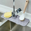 A silicone faucet splash guard mat with a metal dispenser and a wooden brush is placed in a kitchen sink.