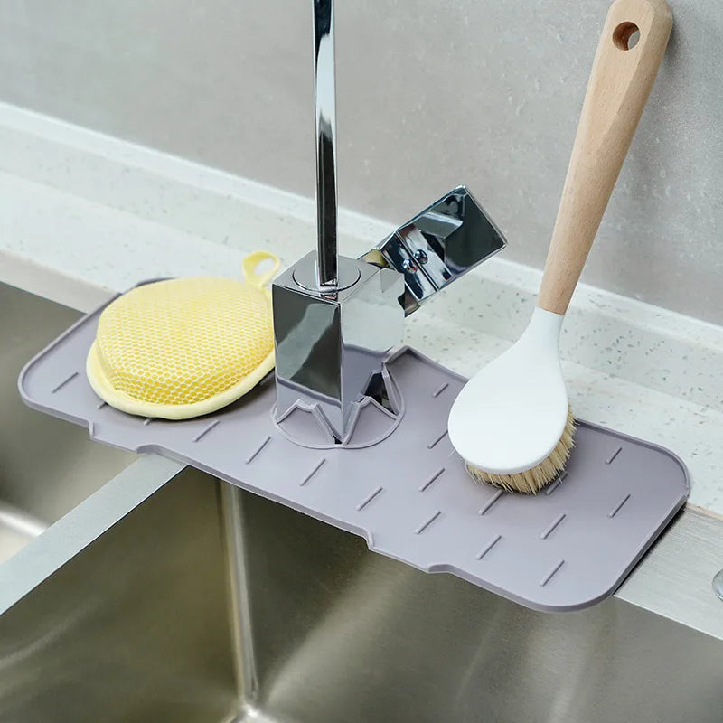 A silicone faucet splash guard mat with a metal dispenser and a wooden brush is placed in a kitchen sink.