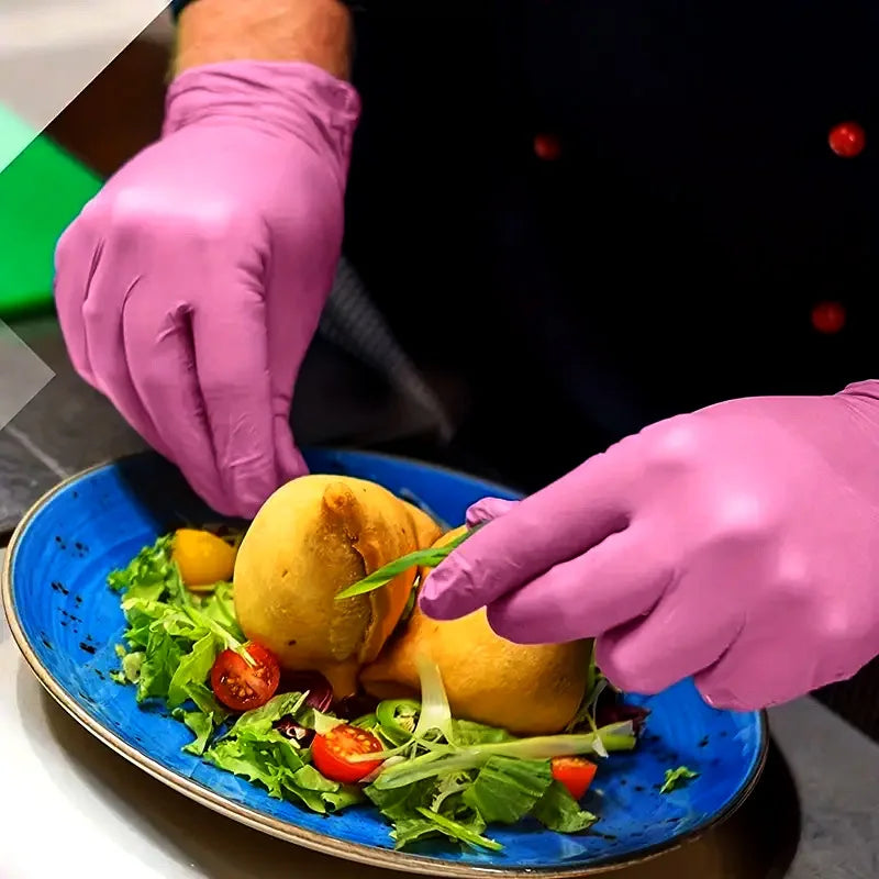 Una mano con un guante desechable rosa de nitrilo está colocando comida sobre una ensalada en un tazón azul.