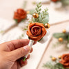Une main tient une boutonnière vintage en soie orange avec une rose et de la verdure.