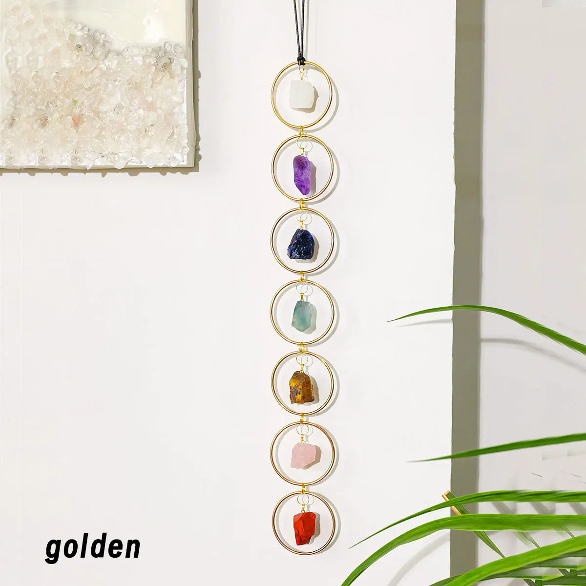 A vertical arrangement of seven crystal healing ornaments with gold-toned chains hanging against a white wall.