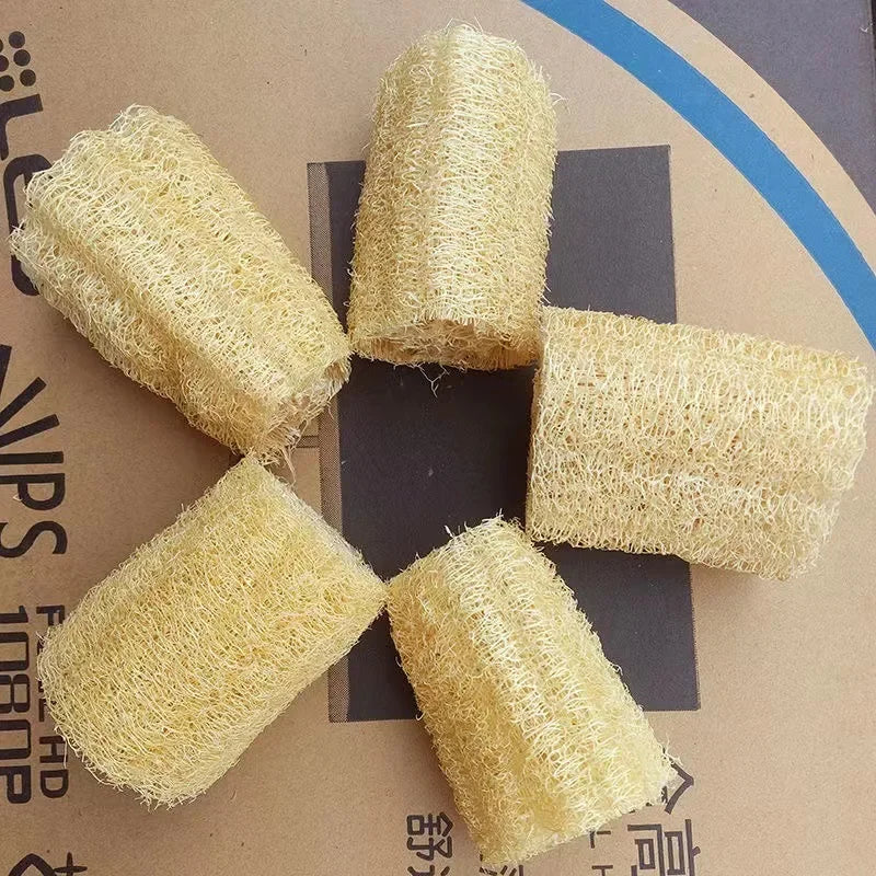 Five cylindrical natural loofah exfoliating body sponges with hanging ropes arranged in a cross pattern.