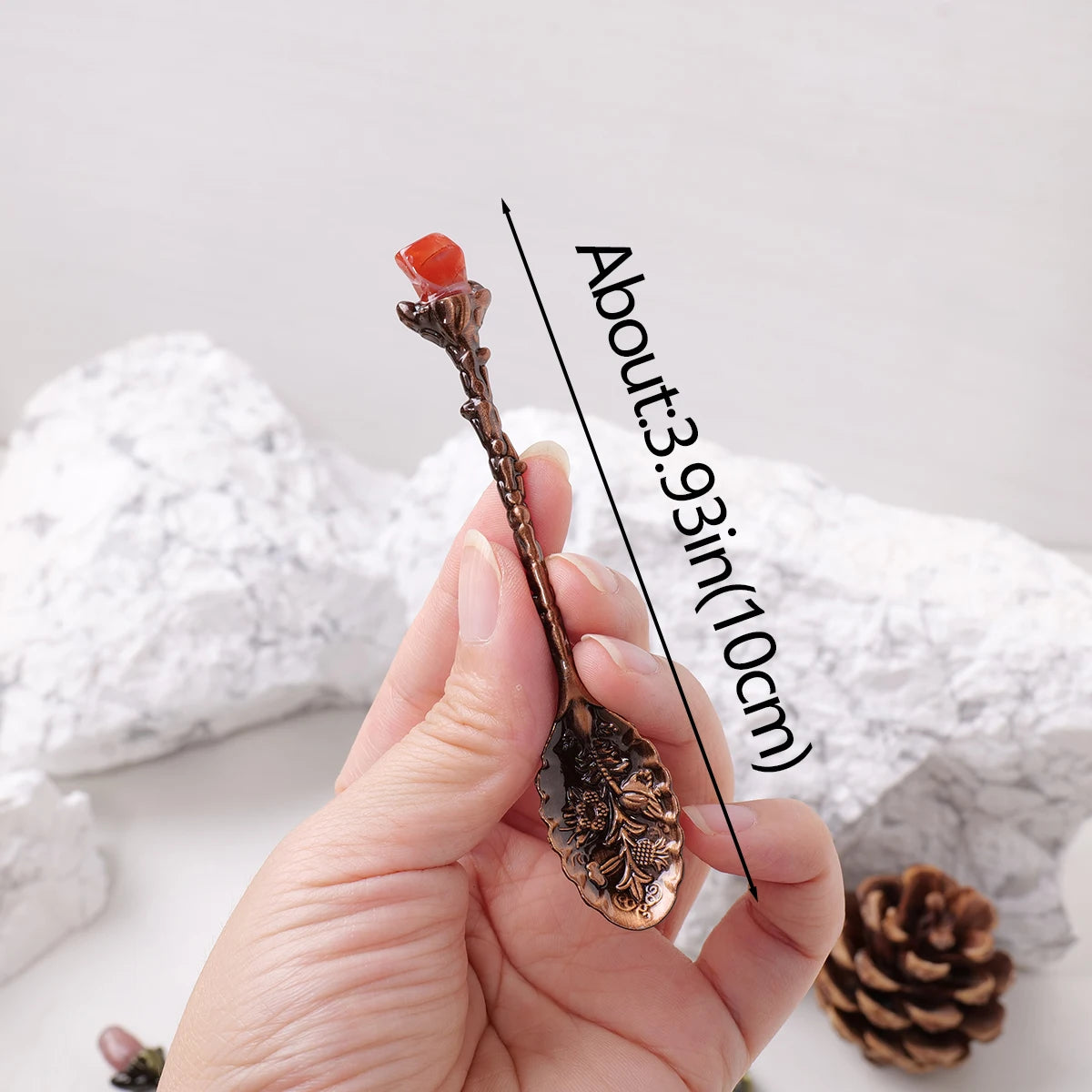 A hand holds a natural crystal spoon with a reddish-orange crystal on the tip against a white background.