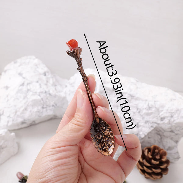 A hand holds a natural crystal spoon with a reddish-orange crystal on the tip against a white background.