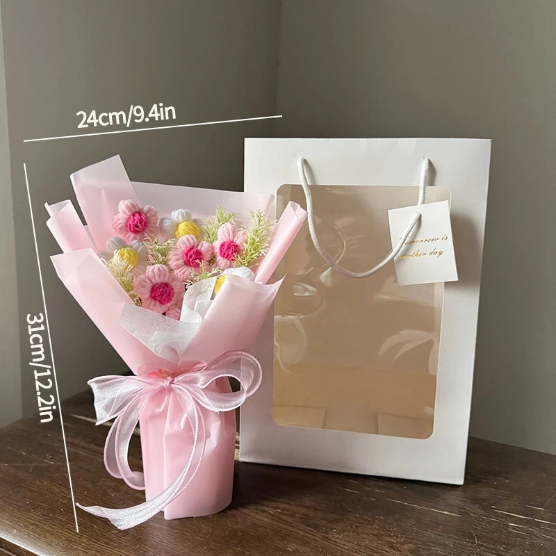 A pink handmade crochet flower bouquet displayed in a white paper bag with a gold string handle.