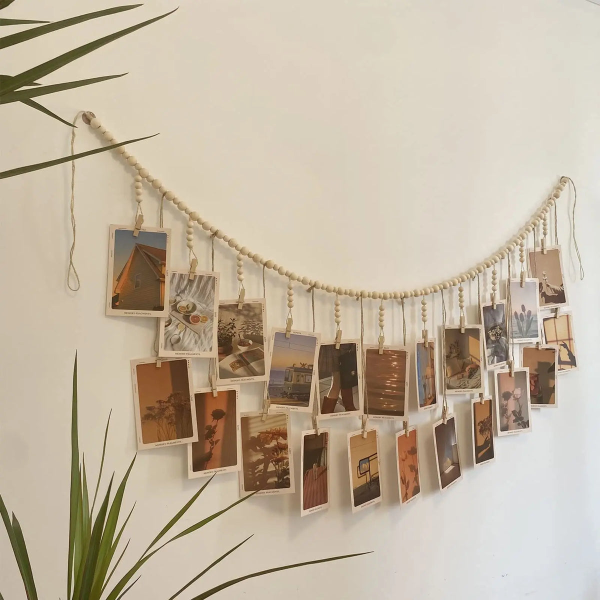 Guirnalda de cuentas de madera bohemia con fotos enmarcadas suspendidas en hilo contra una pared blanca.