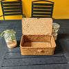A light brown handwoven rattan storage basket with a lid is displayed on a dark wooden table.
