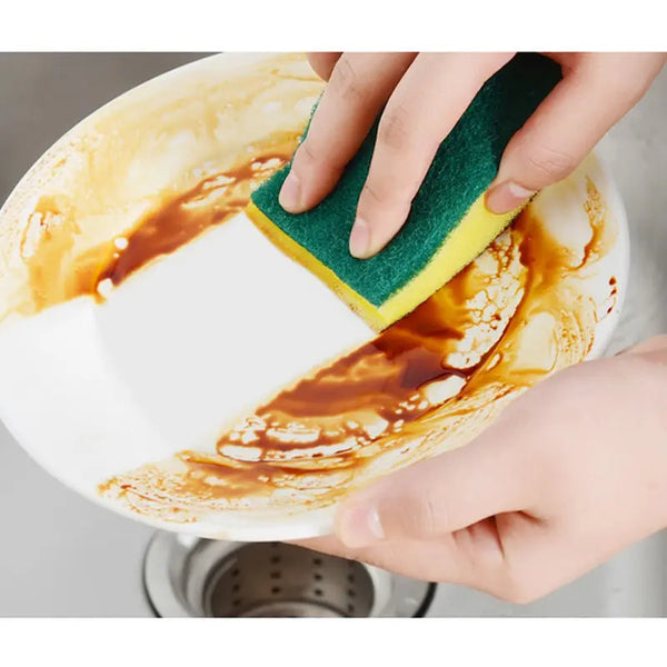 A hand is using a green and yellow double-sided scouring sponge to clean a white ceramic plate stained with orange food resid