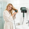 Une femme blonde en robe de chambre utilise un porte-sèche-cheveux fixé au mur au-dessus d'un évier.
