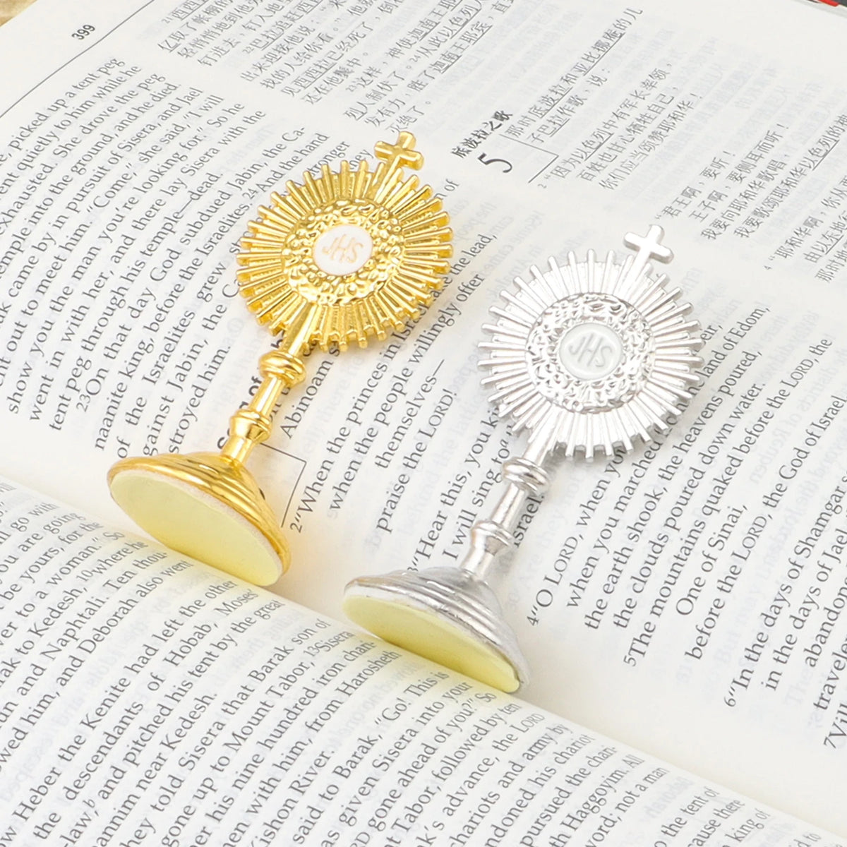 A gold-colored mini metal religious ornament featuring a cross and IHS monogram is displayed on a piece of paper.