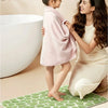 A young girl stands on a green textured bathroom mat next to an adult woman who is holding a pink towel near a white bathtub.