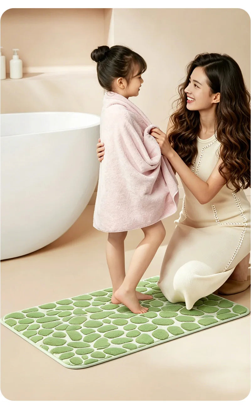A young girl stands on a green textured bathroom mat next to an adult woman who is holding a pink towel near a white bathtub.