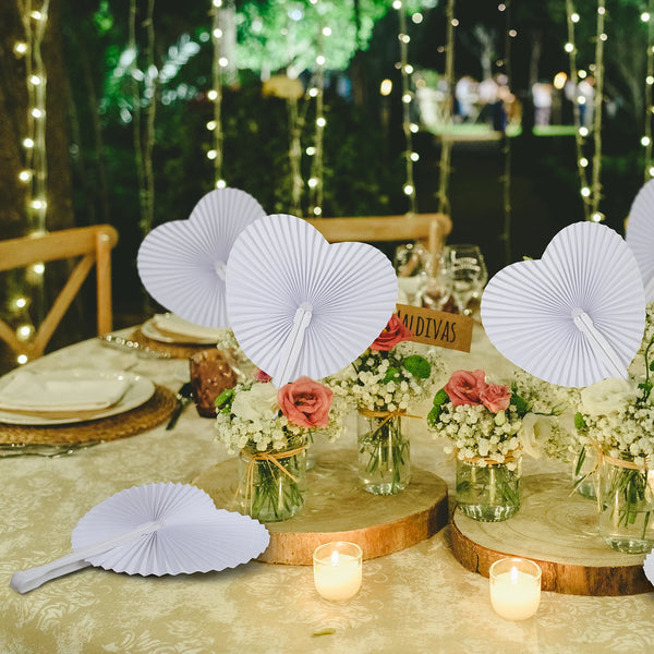Round white folding paper hand fans displayed on a table setting with greenery and string lights.