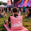 A person sits on a pink mat outdoors with a pink laundry bag and a water bottle.