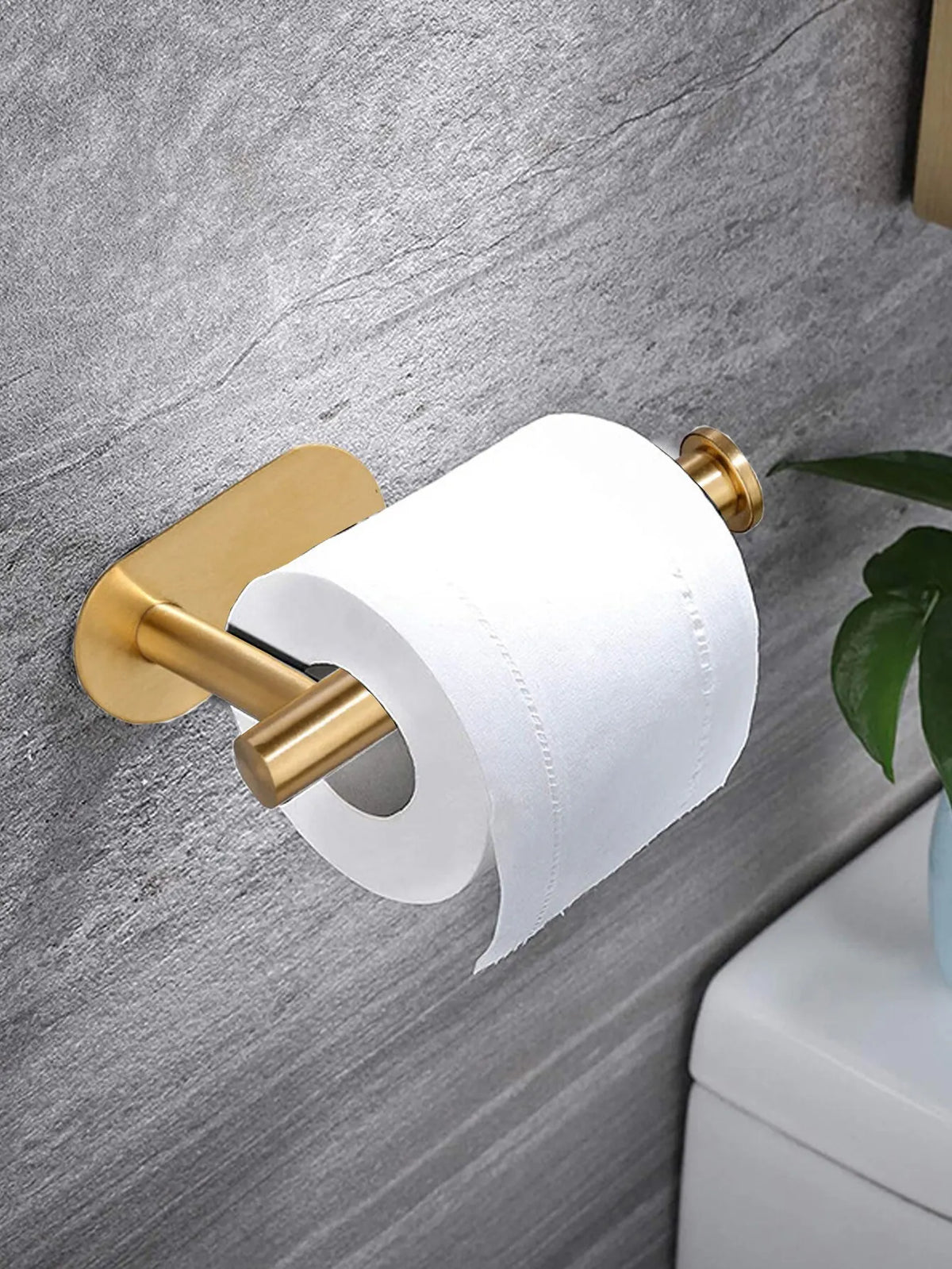 A gold-toned no-drill adhesive paper towel holder with a white roll mounted on a gray wall.