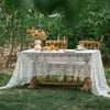 A rustic outdoor table set with a lace tablecloth, wooden chairs, and floral arrangements in a garden setting.