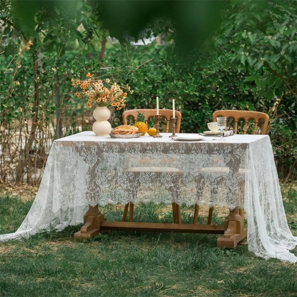 A rustic outdoor table set with a lace tablecloth, wooden chairs, and floral arrangements in a garden setting.