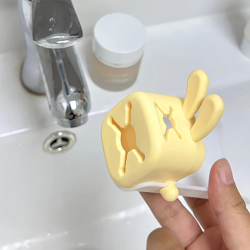 A yellow silicone faucet extender shaped like a cute rabbit is held by a hand next to a glass and a white bottle.