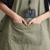 A person's hands are placing a leather pouch into the pocket of a sage green waterproof outdoor work apron.