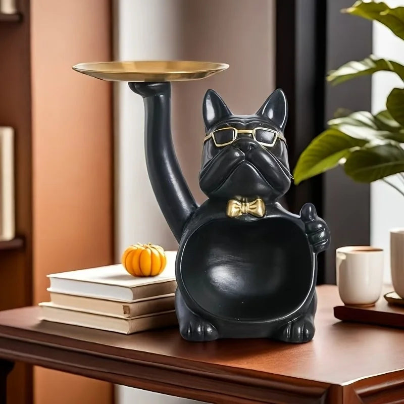 A black French bulldog sculpture holding a gold coin bank and a small snack holder on a wooden table.