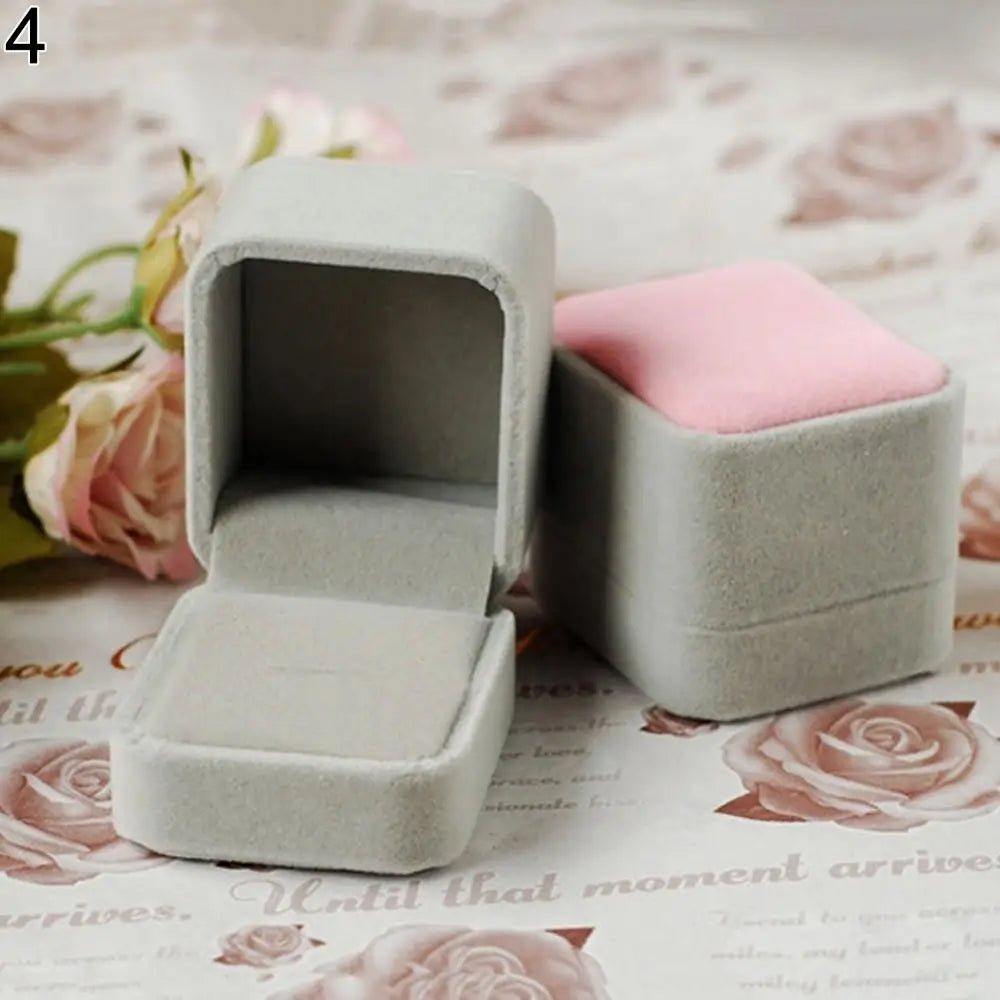 A light gray velvet jewelry box is shown open, with a pink insert, resting on a floral patterned surface.