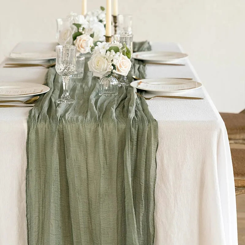 A linen table setting featuring a sage green cheesecloth runner, white plates, silverware, and floral centerpieces.