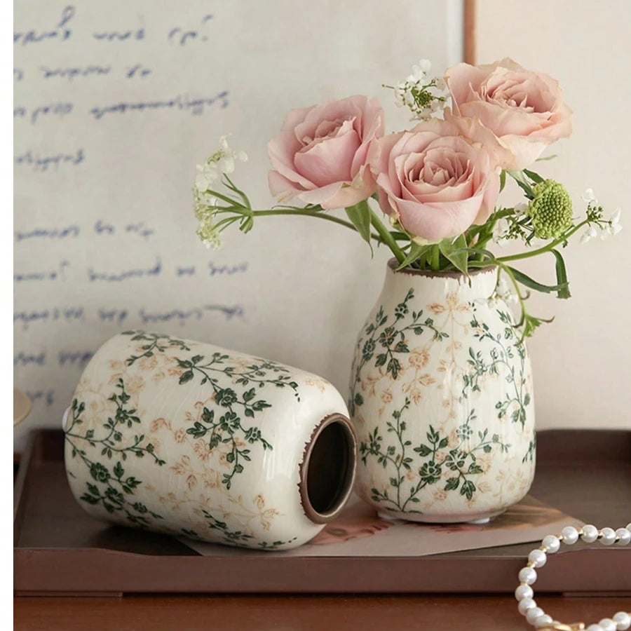 A ceramic bud vase with a floral pattern holds pink roses and green foliage.