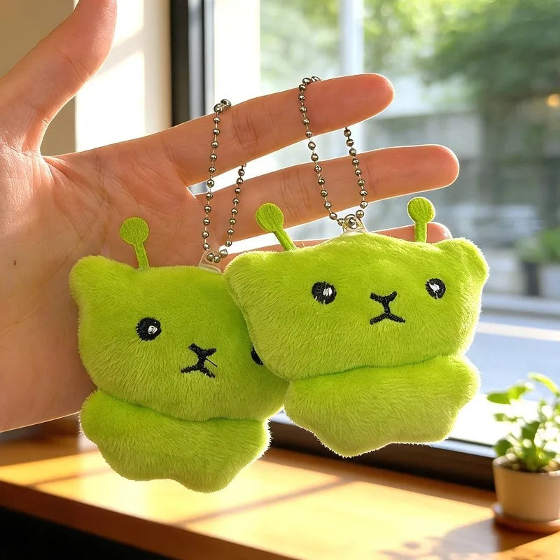 Two bright lime green plush cat keychains are held in a hand against a blurred window background.