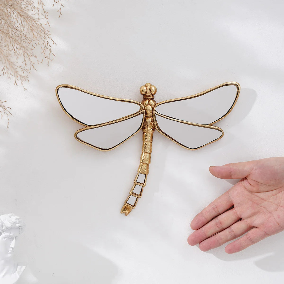 A hand holds a golden dragonfly mirror wall decor featuring translucent wings and a slender body.