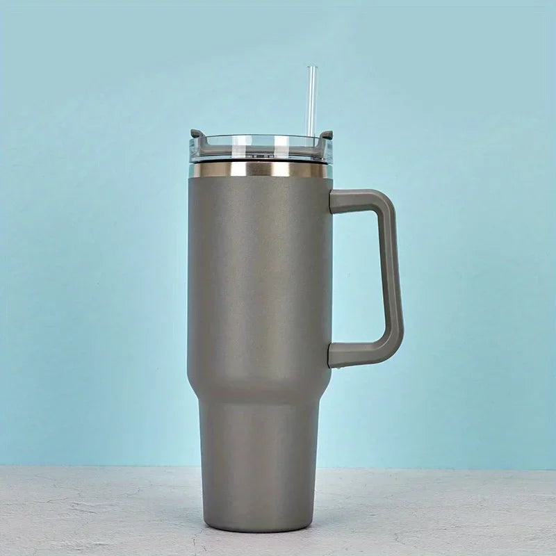A silver insulated stainless steel tumbler with a handle and straw standing on a light surface.