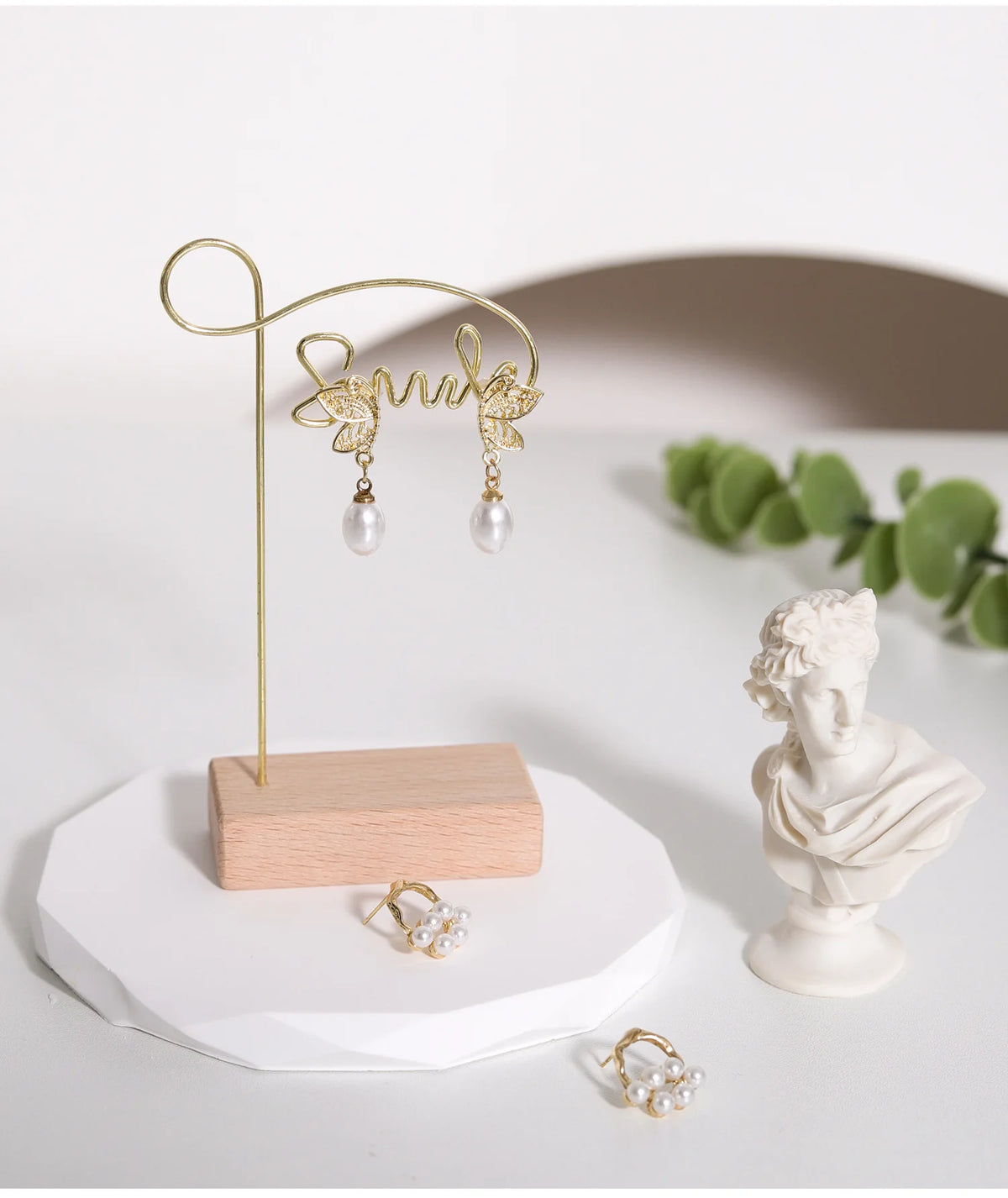 A gold wooden jewelry display rack with a cursive initial hanger and pearl earrings displayed on a white surface.