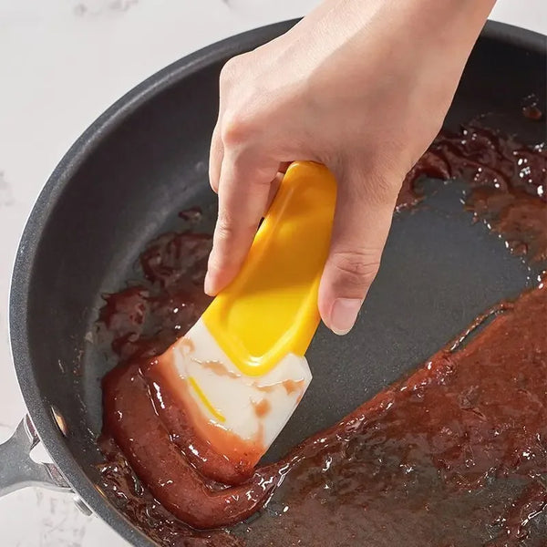 A hand uses a yellow silicone spatula to stir a brown mixture in a dark frying pan.